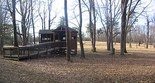 An open wooden structure with a ramp leading up to it, on the edge of a large open field with large leafless trees around