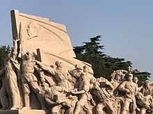 A brown stone sculpture of soldiers fighting with various weapons, led by one carrying a flag with a man's face in profile on it