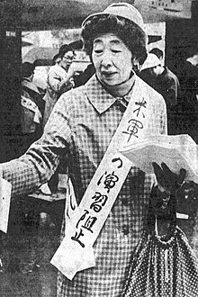 Portrait of an Asian woman in a hat wearing a checked coat draped with a sash bearing a Japanese slogan on her left shoulder and carrying a large bag in her left hand.