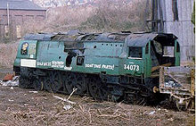 A side-and-rear view of a large 4-6-2 steam locomotive with flat metal side sheets, although some fittings are missing. It is without a tender and stored awaiting purchase for restoration.