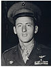 Head and shoulders of a young white man with a peaked cap pushed high up on his forehead, wearing a military jacket with two rows of ribbon bars on the left breast and a star-shaped medal hanging from a ribbon around his neck.