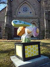 Three-foot high sculpture of a bee, painted in soft colors and with poetic excerpts, on a plinth in front of a stone church.