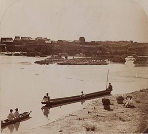 historic black-and-white image of a wide river with rectangular thatch houses on the right bank and several long dugout canoes in the foreground