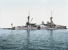 A large gray battleship with two tall masts sits idly in calm waters. Three small boats are tied alongside.