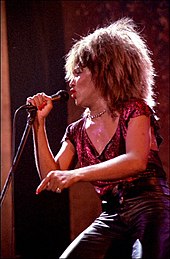 A woman holding onto a microphone and wearing a necklace, a sparkly red-colored top, and leather pants.