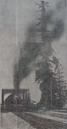Grainy image of train crossing trestle, belching black smoke onto the tall tree