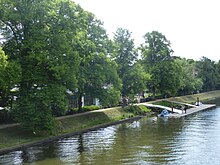 Zu sehen ist der Kanal vorn, ein grüner Uferstreifen und dahinter ein von Bäumen gesäumter Weg. Das Bootshaus ist weitgehend von den Bäumen verdeckt, während der Steg gut erkennbar ist.