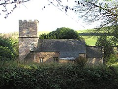Stone building with tiled roof and square tower, surrounded by vegetation.