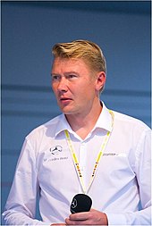 Head and shoulders of a man in his forties with blonde hair and grey eyes. He is wearing a white shirt which bears the Mercedes-Benz and AMG logos, and is holding a microphone in both of his hands.
