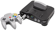 The Nintendo 64 controller is the light gray controller with three handles for the player's two hands. It has red, green, blue, and yellow buttons, an analog stick, and a directional pad. The controller is plugged into the charcoal gray Nintendo 64 with a light gray cartridge inserted. The sleek console is convex on its top and has two power switches and four controller ports.