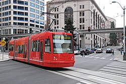 A red streetcar on a single track crosses a city street, with cars stopped in the background under a monorail track.