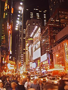 Photograph of a crowded city street at night. The street is in a commercial district; the buildings all have at least several stories, and some are high rise. The buildings have elaborate signs, many of which incorporate neon lighting. There are prominent signs for Madame Tussaud's, Loew's, Empire, AMC 25 Theatre, and Modell's.