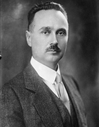 A man with combed-back hair and a small mustache wearing a three-piece tweed suit.