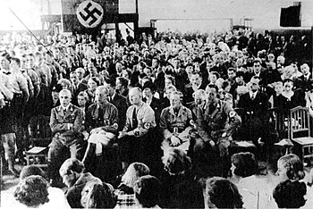 Black-and-white photograph of a large assembly with a Nazi flag on the back wall. Hitler Youth stand on the left. Seated in the front row are suited men with Nazi armbands; the central baldish man has a toothbrush moustache. Behind him are a hatted woman and a man with no sideburns, flanked by four-plus children.