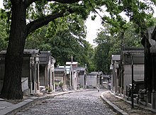 A cobbled street stretches out from the foreground and bends to the left. The street is lined with above-ground tombs, and a number of trees appear in the background.