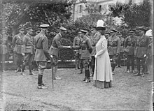 A black-and-white photograph showing a uniformed man on the left who is stooping slightly forward as he holds the hand of a man on the right, who is the king. Also in the photograph are many other uniformed men, forming a group mostly behind the two main subjects. A woman wearing a feathered hat stands to the left side of the king.