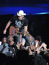 A man in a dark t-shirt, jeans and cowboy hat playing the guitar on a stage in front of an audience