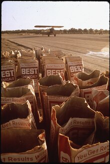 Rows of opened pesticide bags are lined up against a field with a pesticide distributor plane in the background. The pesticide bags read 'You get results with Best Chemicals".