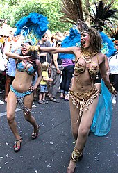 Frontale Farbfotografie von zwei Sambatänzerinnen, die mit weiten Armbewegungen und hellblauen Kostümen durch eine Straße laufen. Im Hintergrund ist eine Menschenmenge.