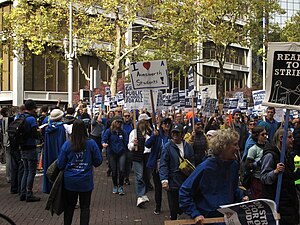 People march through a city street, wearing blue and carrying signs: "I heart Ainsworth students," "great public schools for all," "PAT on strike," among others. On the left, musicians play, facing the marchers.