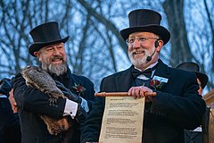 alt=Two smiling men dressed in formal attire stand beside each other. Other people and bare trees are visible in the background. The man on the left holds a groundhog in his arms. The man on the right, wearing an earpiece, holds forward an open scroll, which reads in small text: "HEAR YE, HEAR YE, HEAR YE. TODAY, 2-2-22, WELCOME TO PUNXSUTAWNEY TO CELEBRATE GROUNDHOG DAY, THE ONE HUNDRED THIRTY SIXTH ANNUAL TREK OF THE PUNXSUTAWNEY GROUNDHOG CLUB. PUNXSUTAWNEY PHIL, THE SEER OF SEERS, THE PROGNOSTICATOR OF ALL PROGNOSTICATORS, WAS GENTLY LIFTED FROM HIS BURROW AT 7:25 AM, AND HELD HIGH TO SEE. HIS FAITHFUL FOLLOWERS HAD RETURNED WITH GLEE! PLACING PHIL ON TOP OF THE STUMP, WHERE IN GROUNDHOGESE, HE DIRECTED THE PRESIDENT, JEFF LUNDY, AND THE INNER CIRCLE TO HIS PREDICTION SCROLL THAT READS: WINTER HAS BEEN BLEAK AND DARK AND BEREFT OF HOPE. YET WINTER IS JUST ANOTHER STEP IN THE CYCLE OF LIFE. AS I LOOK OUT OVER THE FACES OF THE TRUE BELIEVERS FROM AROUND THE WORLD, I BASK IN THE WARMTH OF YOUR HEARTS. I COULDN'T IMAGINE A BETTER FATE, WITH MY SHADOW I HAVE CAST, THAN A LONG AND LUSTROUS SIX MORE WEEKS OF WINTER."