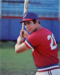 A man wearing a red baseball jersey with blue and white trim and a white "20" on the back with a blue cap poses holding a baseball bat with both hands as if ready to swing.