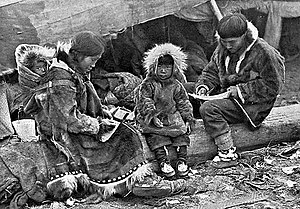 An Inuit family is sitting on a log outside their tent. The parents, wearing warm clothing made of animal skins, are engaged in domestic tasks. Between them sits a toddler, also in skin clothes, staring at the camera. On the mother's back is a baby in a papoose.