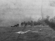 A large ship is sinking in the distance; a large dense cloud of smoke emanates from the wreck.