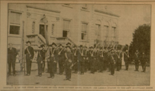 "Black and white photograph of a large group of men in military uniform, many carrying rifles, pictured outside a building, with James Larkin in civilian clothes standing to the left"