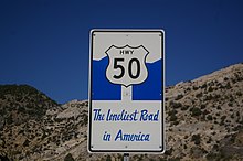 rectangular sign with an image of a retro Highway 50 sign against a stylized blue mountain range with the words, "The Loneliest Road in America" written in a cursive font
