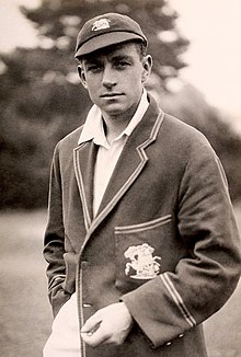 A dark-haired man in a cricket blazer and cap looks at the camera