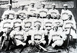 Baseball players are posing for a photograph, six men standing, seven men sitting on chairs, and three are sitting on the ground.