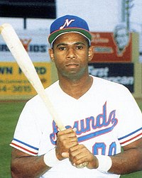 A man wearing a white baseball uniform with "Sounds" on the chest in blue and red and a blue cap with a white "N" on the center poses holding a baseball bat with both hands.