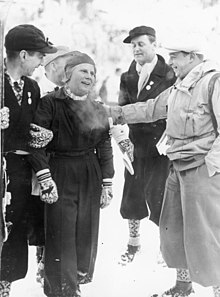 A Scandinavian woman is pictured standing surrounded by three men, one of whom is seen patting her shoulder.