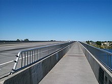 A bridge with metal railings seen from a wide pedestrian path that abuts a freeway