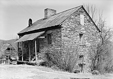 A three story stone building with peaked roof and a front porch to the left built at the crest of a depression to the right