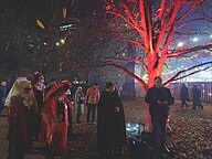 A group of people stood next to a tree lit in red; the tree is bare, with leaves on the ground