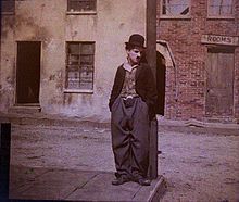 Frontale Farbfotografie eines Mannes, der an einem Pfahl an der Straßenecke lehnt. Er trägt einen Melonenhut, einen kurzen Schnurrbart, eine Weste und weite Hosen. Sein Blick geht nach rechts unten. Im Hintergrund sind heruntergekommene Fassaden.