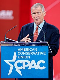 Oliver North speaking from notes behind a red background at a blue podium