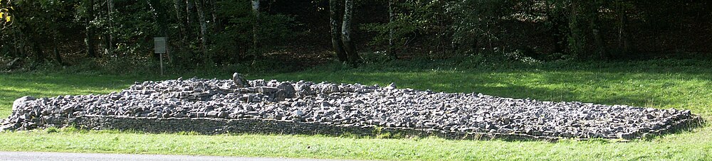 Elongated, elevated view of the cromlech from its side, with the edge of the woods to its rear. The tumulus' trapezium shape is evident, its boulders retained by a short wall, missing at the very front, left, where rubble has tumbled out.