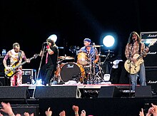 A band performs on a stage while several audience members raise their arms up. In the background, two spotlights flash brightly.