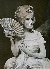 Black and white photo of a white woman in a wig with a fan.