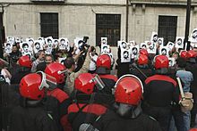 Demonstrators holding pictures in front of uniformed people in red helmets, perhaps police officers