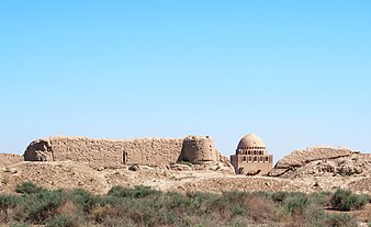 Ruined walls rise out of small spiky bushes under a cloudless sky; in a gap can be seen a mosque-like structure, with a camel providing a sense of scale.