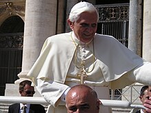 Frontale Farbfotografie von einem Papst mit einem goldenen Kreuz als Halskette, der sich an einem weißen Geländer festhält und seinen linken Arm lächelnd aus dem Foto streckt. Weiter unten und um ihn verteilt stehen Männer in schwarzen Anzügen.