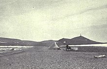 A conical tent stands on a desolate stretch of beach, with a range of low hills in the background.