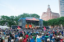 A large crowd is gathered in front of an outdoor stage with an orchestra and singer performing. The skyline of New Haven rises in the background behind the colorful stage.