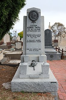 granite grave with headstone. a brass relief showing the head of Albert Jack is at the top of the headstone. A soldier's slouch hat and sword made of brass lay on top of the grave.