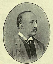 A cameo engraving of a balding mustachioed man in profile view looking to the viewer's right. The man is dressed in a three-piece suit with a high-cut waistcoat and a combination of a wing collar and a necktie.