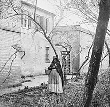 A black and white photo of a Black woman standing in front of a dilapidated and overgrown building.